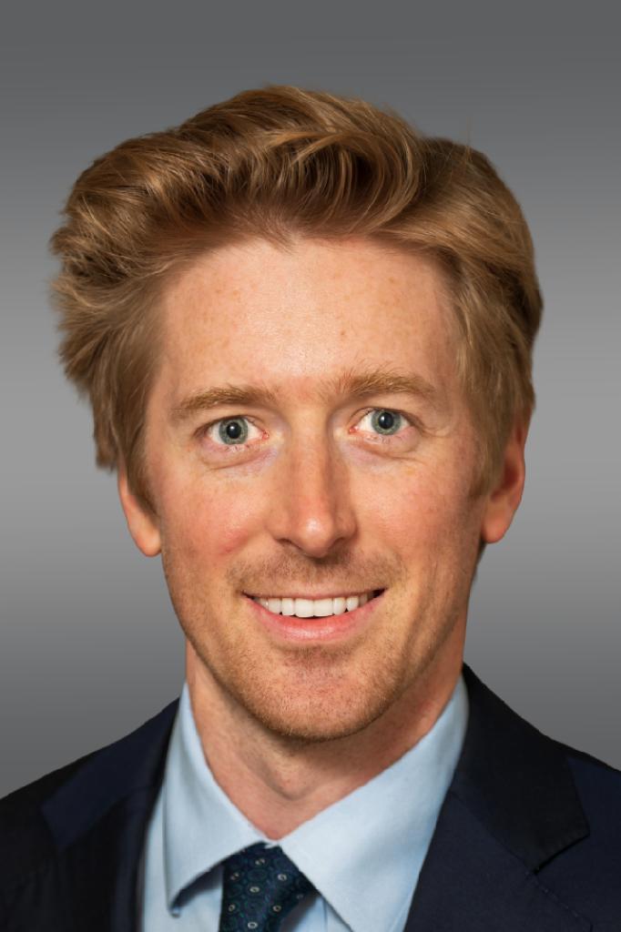 Head-and-shoulders portrait of a smiling man in a navy suit and light blue shirt against a gray gradient background.